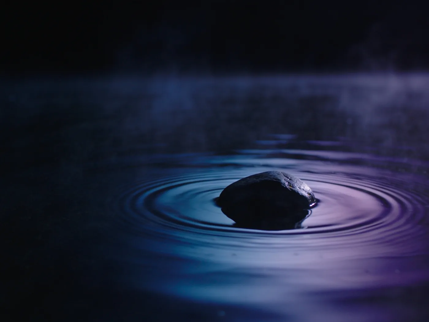 Ein dunkler Stein in völlig stillem Wasser bei Nacht, ein einzelner Wellenring breitet sich aus, leichter Nebel.